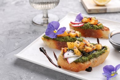 Photo of Delicious bruschettas with pesto sauce, tomatoes, balsamic vinegar and violet flowers on gray table, closeup
