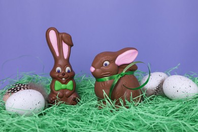 Photo of Easter celebration. Funny chocolate bunnies and painted eggs with feathers on grass against violet background