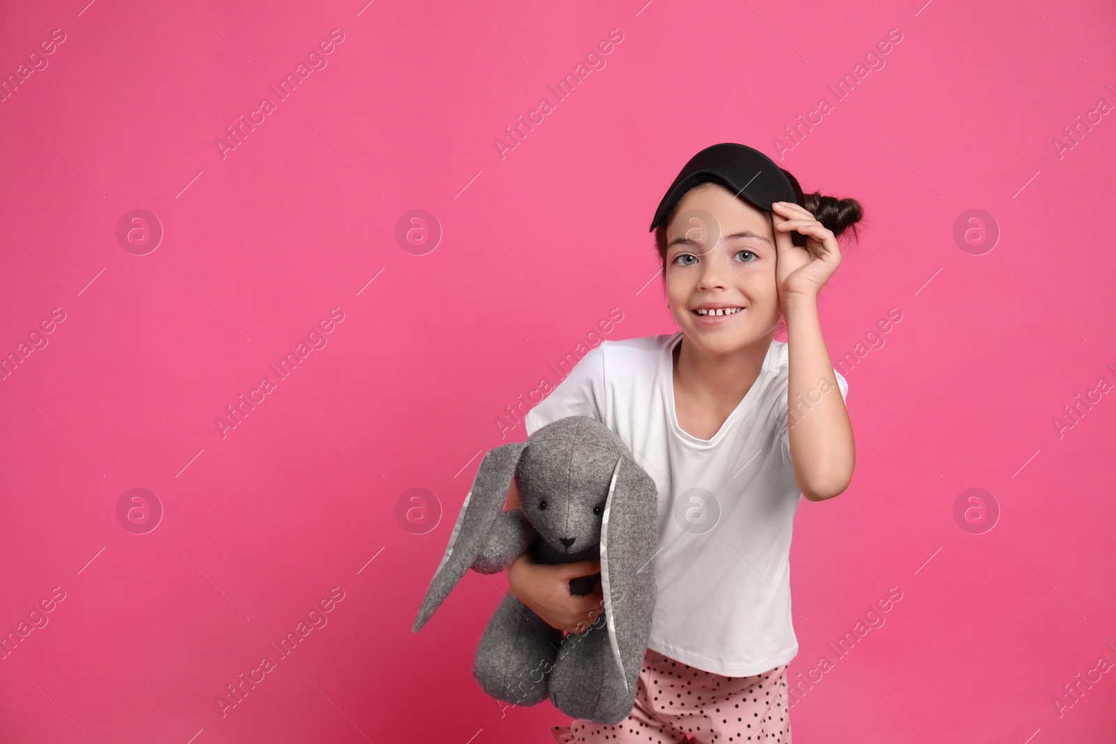 Photo of Cute girl in pajamas with sleep mask and toy bunny on pink background. Space for text