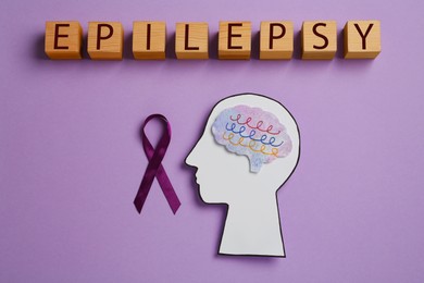 Photo of Human head cutout with brain, purple ribbon near word Epilepsy made of wooden cubes on lilac background, flat lay
