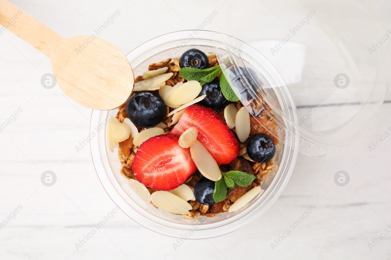 Photo of Tasty granola with berries, almond flakes and mint in plastic cup on white wooden table, top view