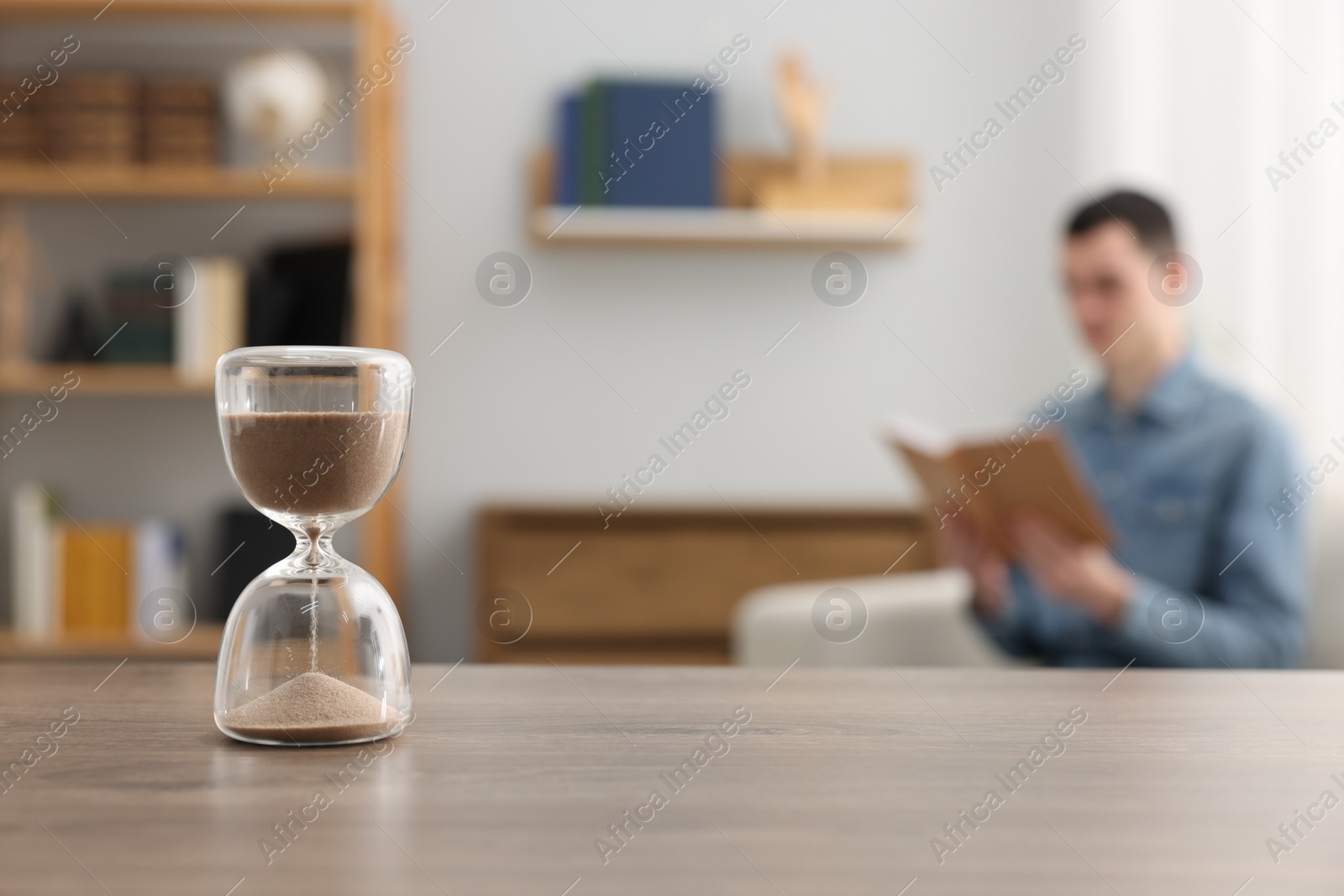 Photo of Hourglass with flowing sand on desk. Man reading book in room, selective focus