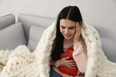 Photo of Woman using hot water bottle to relieve abdominal pain on sofa at home