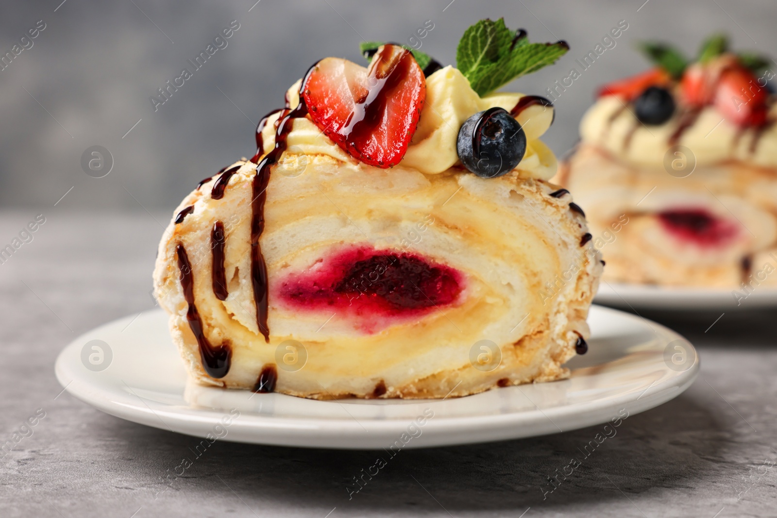 Photo of Piece of tasty meringue roll with jam, cream, strawberry, blueberry and mint on light grey table, closeup