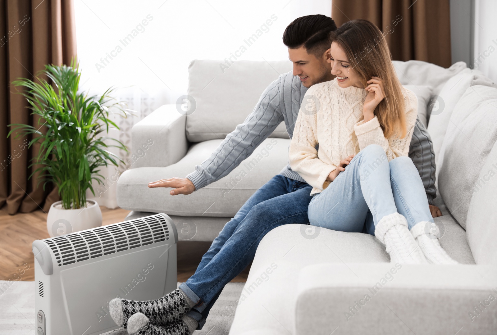 Photo of Happy couple sitting on sofa near electric heater at home