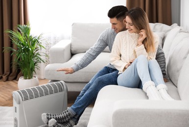 Photo of Happy couple sitting on sofa near electric heater at home