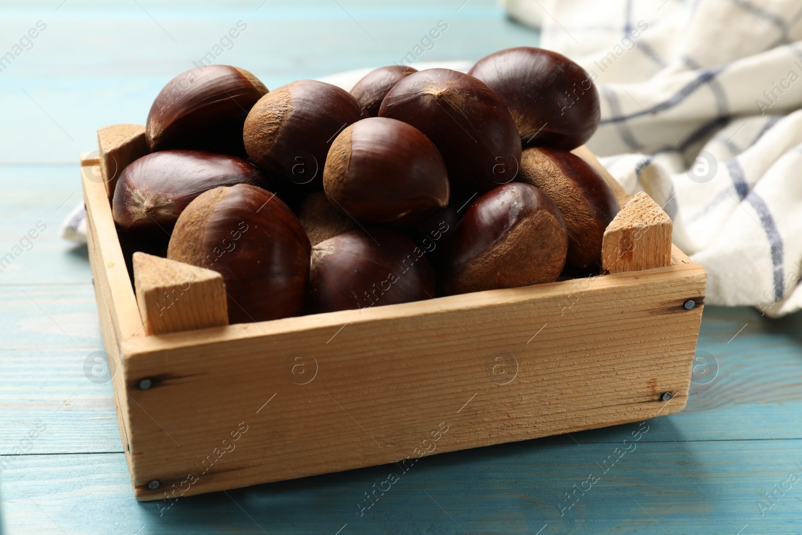 Photo of Roasted edible sweet chestnuts in crate on light blue wooden table, closeup