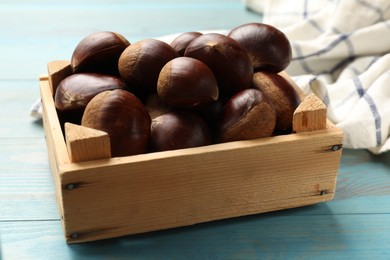 Photo of Roasted edible sweet chestnuts in crate on light blue wooden table, closeup