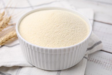 Photo of Uncooked organic semolina in bowl and spikelets on white wooden table, closeup