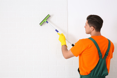 Photo of Professional janitor cleaning white brick wall with squeegee indoors. Space for text