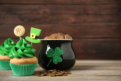 Photo of Decorated cupcakes and pot with gold coins on wooden table, space for text. St. Patrick's Day celebration