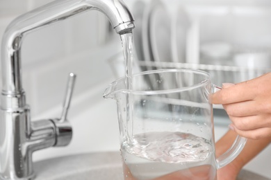 Photo of Woman pouring water into glass jug in kitchen, closeup
