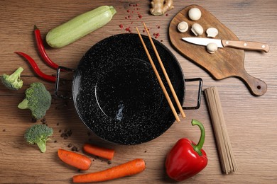 Empty iron wok surrounded by raw ingredients on wooden table, flat lay