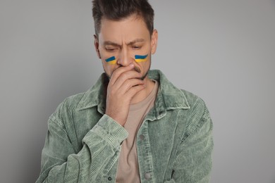 Photo of Sad man with drawings of Ukrainian flag on face against light grey background
