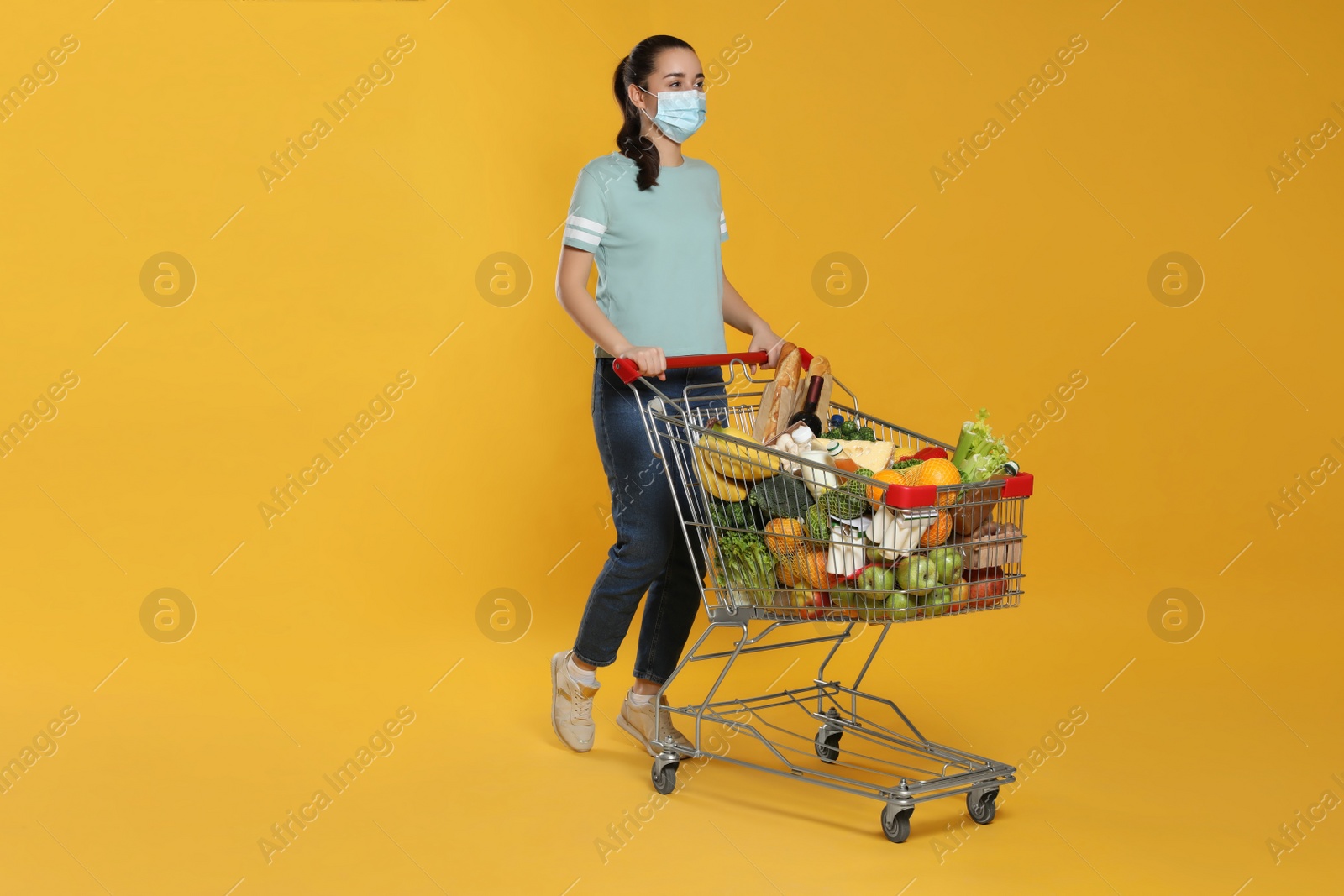 Photo of Woman with protective mask and shopping cart full of groceries on yellow background