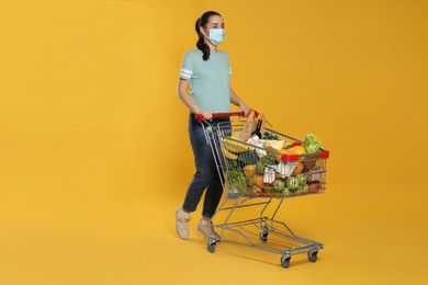 Photo of Woman with protective mask and shopping cart full of groceries on yellow background