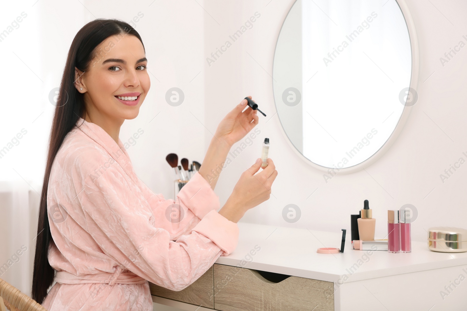 Photo of Beautiful young woman with gel for eyelashes at dressing table indoors