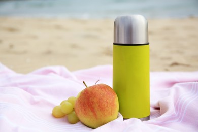 Metallic thermos with hot drink, fruits and plaid on sandy beach near sea