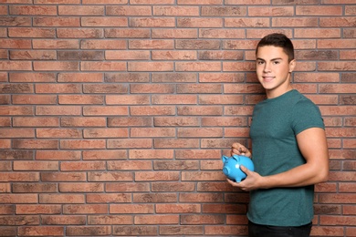 Photo of Teenage boy putting coin into piggy bank near brick wall. Space for text