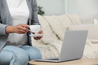 Woman with cup of drink near laptop at wooden coffee table indoors, closeup