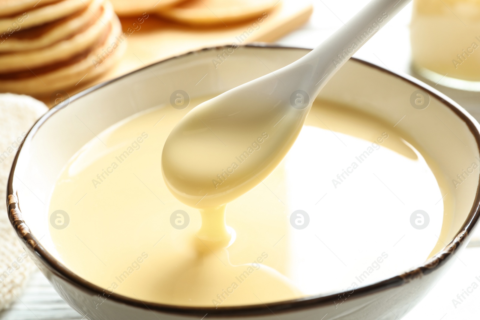 Photo of Spoon of pouring condensed milk over bowl on table, closeup. Dairy products