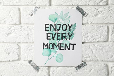 Card with phrase Enjoy Every Moment attached on white brick wall