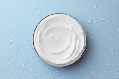 Jar of face cream on light blue surface covered with water drops, top view