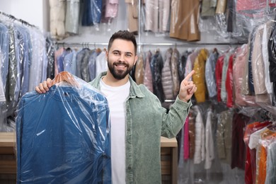 Photo of Dry-cleaning service. Happy man holding hanger with sweatshirt in plastic bag and pointing at something indoors, space for text