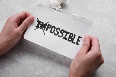 Photo of Motivation concept. Woman holding paper with changed word from Impossible into Possible by crossing over letters I and M at grey table, closeup