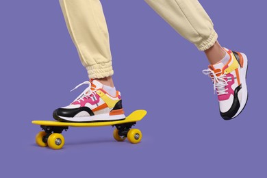 Photo of Woman in new stylish sneakers standing on skateboard against purple background, closeup