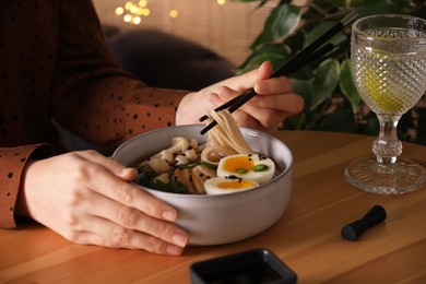 Woman eating delicious ramen with chopsticks at wooden table indoors, closeup. Noodle soup