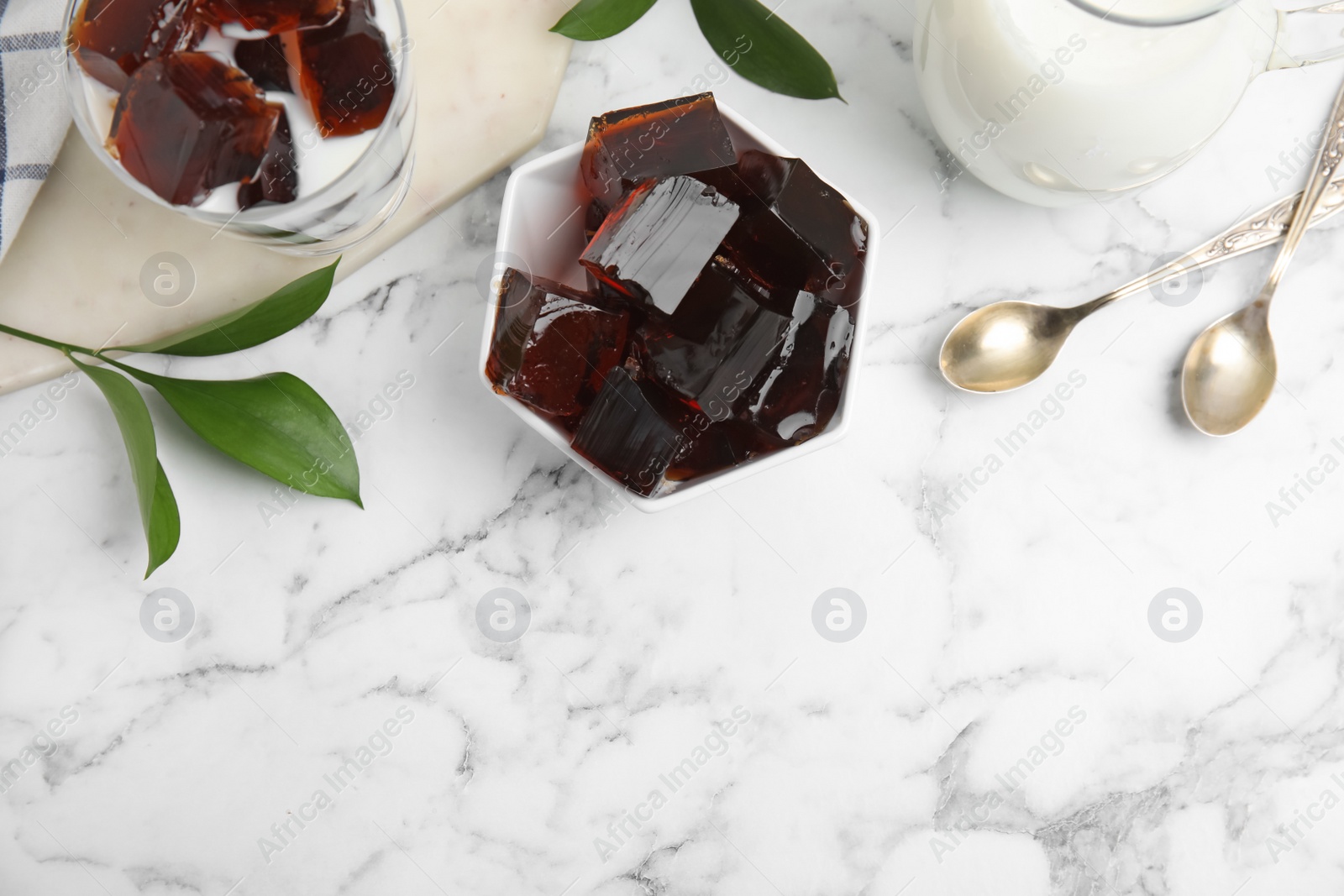 Photo of Delicious grass jelly cubes, milk and green leaves on white marble table, flat lay. Space for text