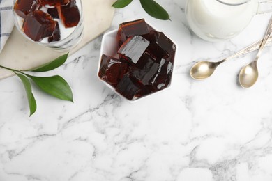 Photo of Delicious grass jelly cubes, milk and green leaves on white marble table, flat lay. Space for text