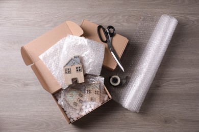 Open box with decorative house figures, adhesive tape, scissors, foam peanuts and bubble wrap on wooden table, flat lay