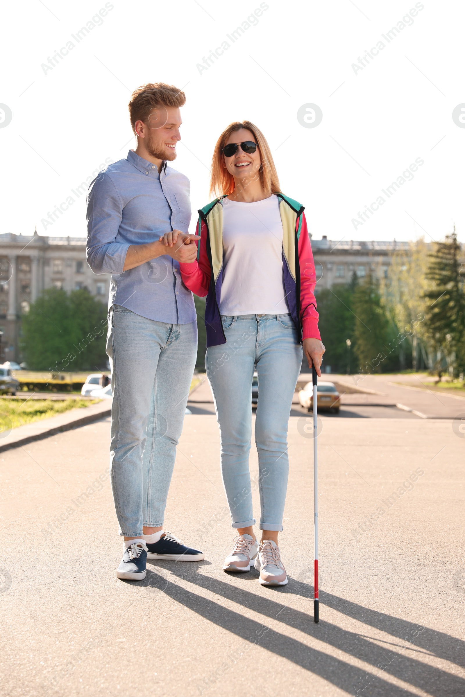 Photo of Young man helping blind person with long cane walking outdoors