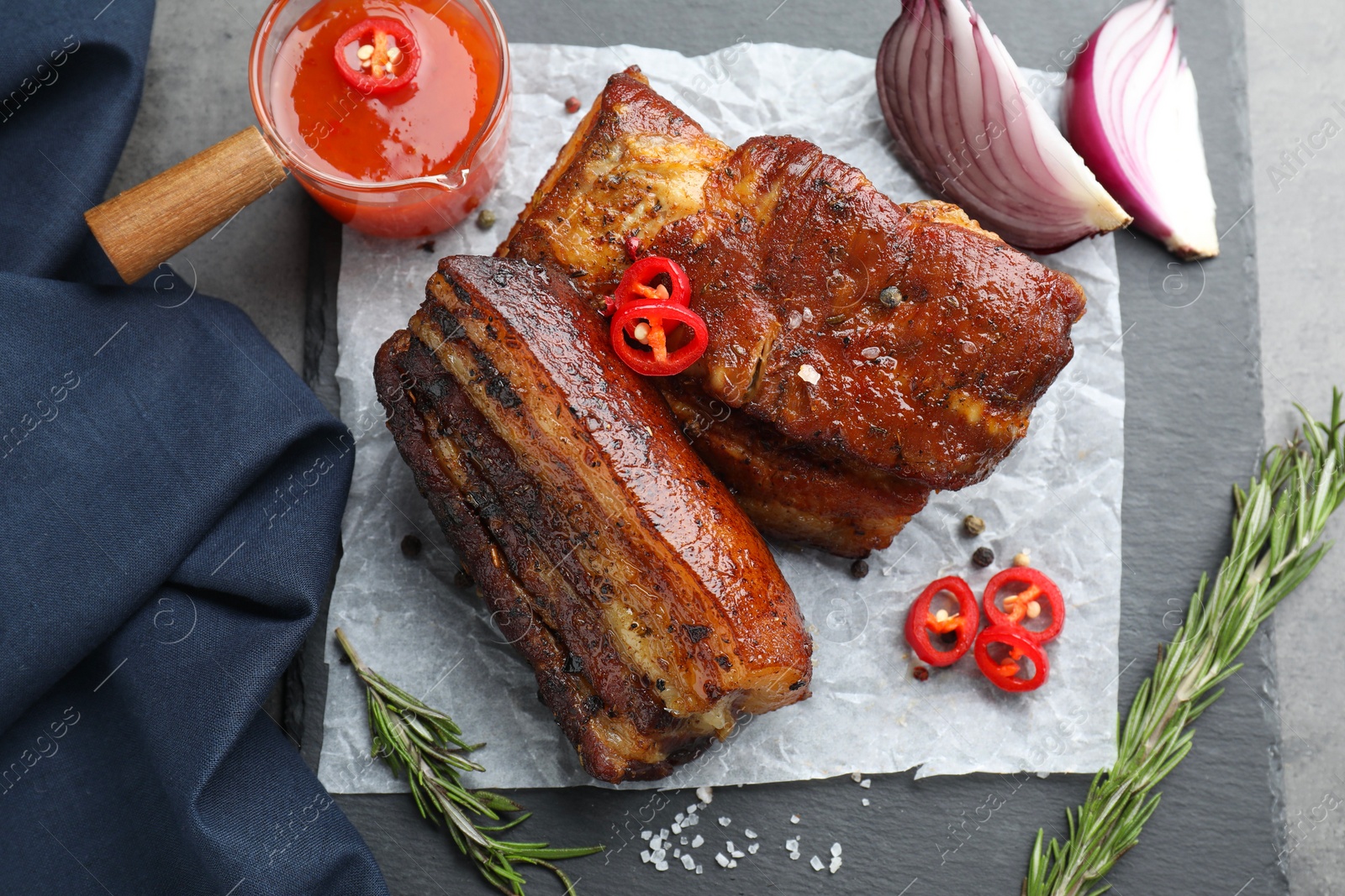 Photo of Pieces of baked pork belly served with sauce, rosemary and chili pepper on grey table, top view