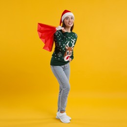 Happy young woman in Christmas sweater and Santa hat with shopping bags on orange background