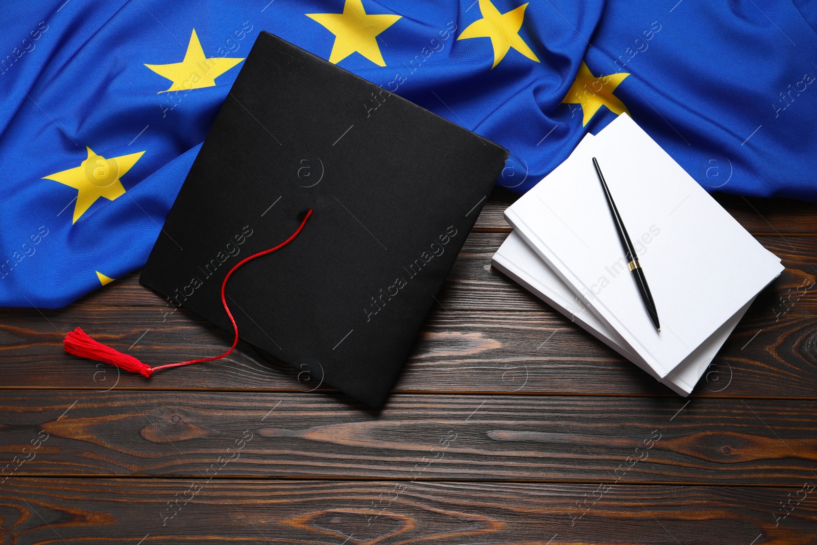 Photo of Black graduation cap, books, pen and flag of European Union on wooden table, flat lay