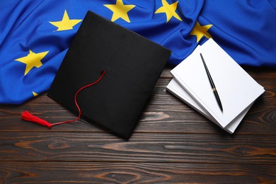 Black graduation cap, books, pen and flag of European Union on wooden table, flat lay