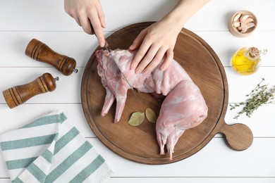 Photo of Woman cutting whole raw rabbit at white wooden table, top view