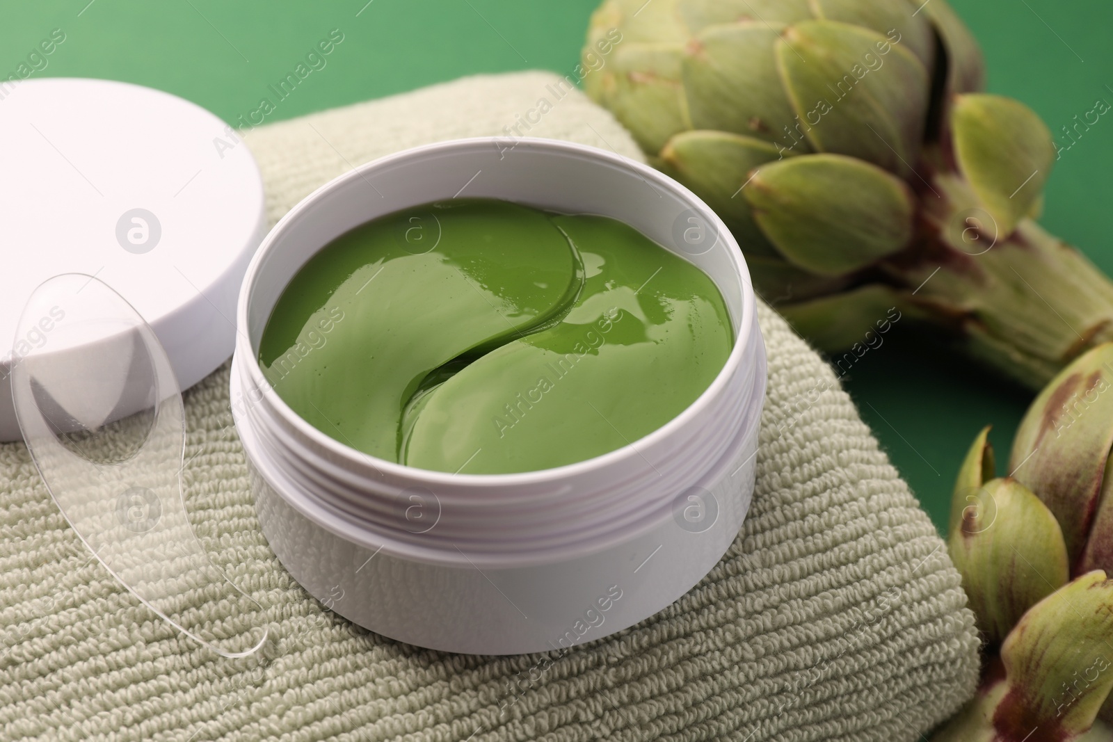 Photo of Package of under eye patches, towel and artichokes on green background, closeup. Cosmetic product