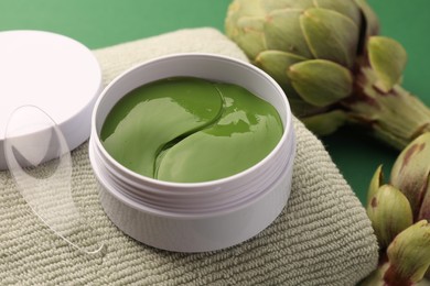 Photo of Package of under eye patches, towel and artichokes on green background, closeup. Cosmetic product