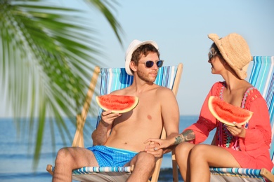 Young couple with watermelon slices in beach chairs at seacoast