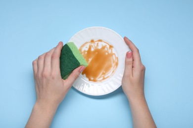 Woman washing dirty plate with sponge on light blue background, top view