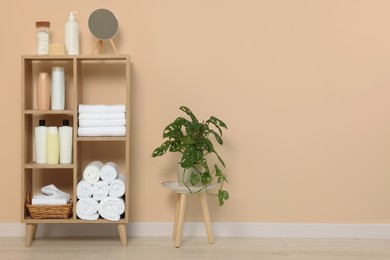 Photo of Soft folded towels and cosmetic bottles on wooden shelving unit near beige wall, space for text