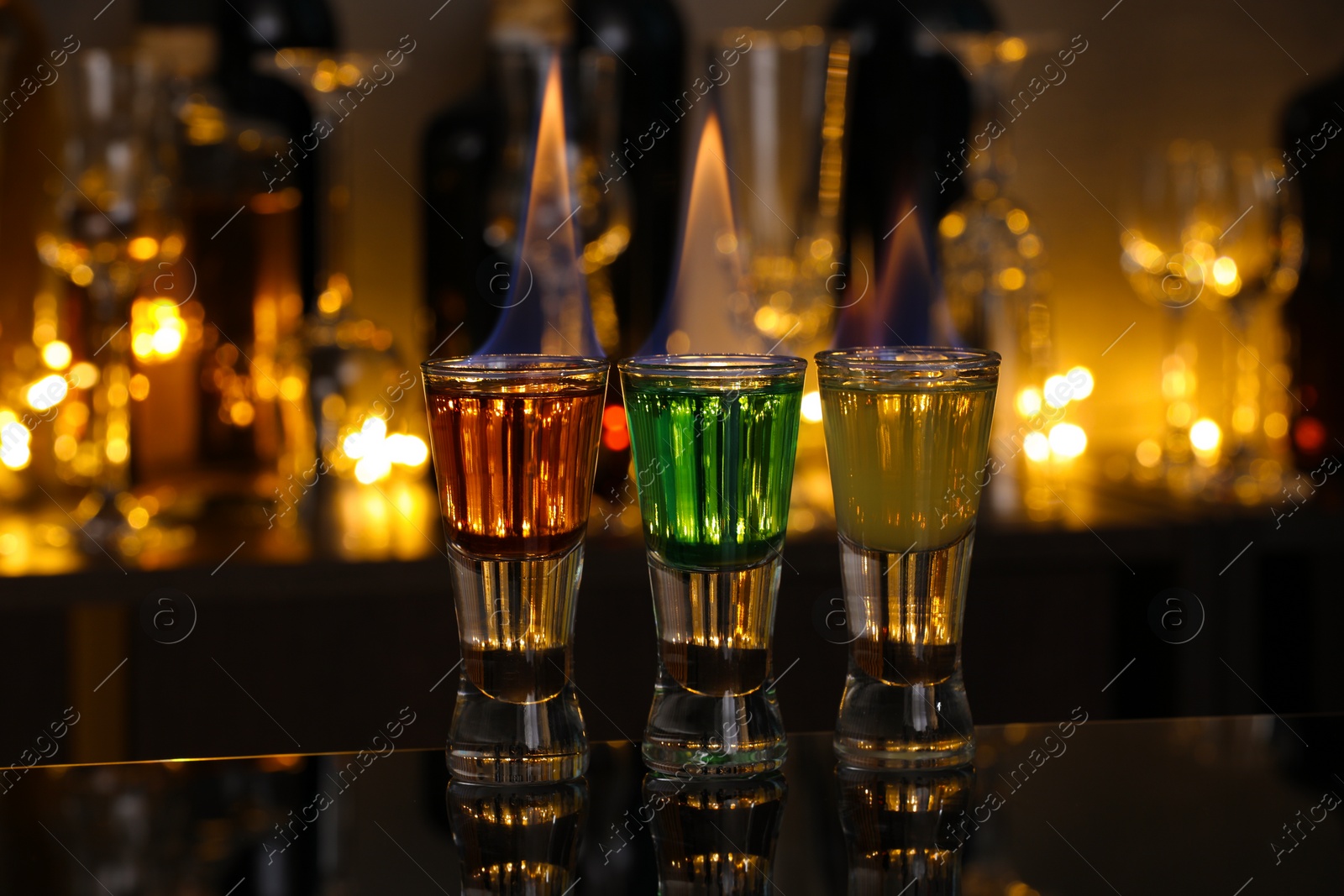 Photo of Flaming shooters in shot glasses on mirror surface against blurred background. Alcohol drink