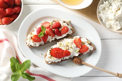 Photo of Delicious ricotta bruschettas with strawberry, mint and pistachios served with honey on white wooden table, flat lay