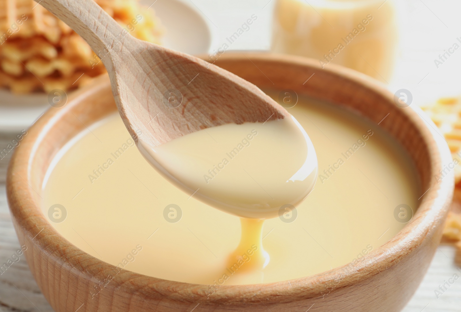 Photo of Spoon of pouring condensed milk over bowl on table, closeup. Dairy products