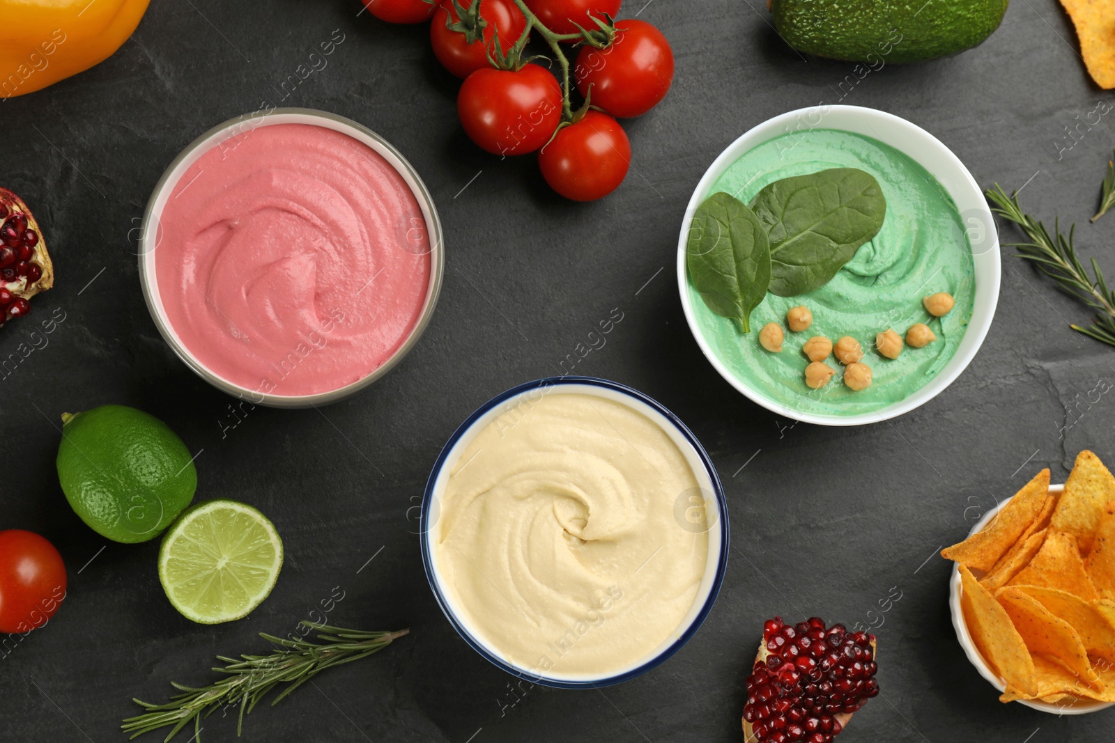 Photo of Different kinds of tasty hummus, nachos and ingredients on black table, flat lay