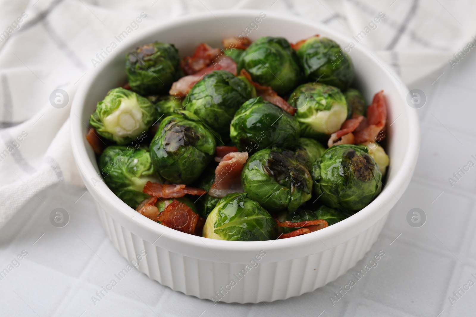 Photo of Delicious roasted Brussels sprouts and bacon in bowl on white tiled table, closeup